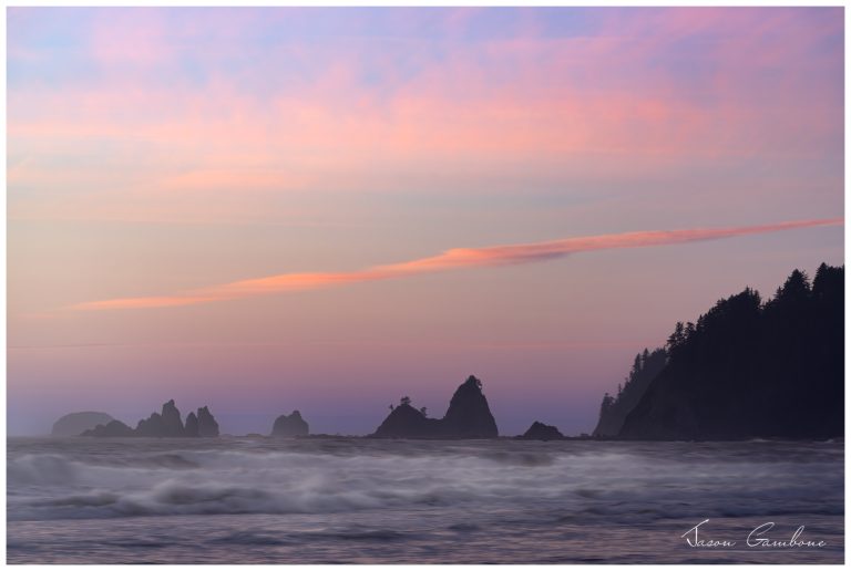 Pastel sunset over Rialto Beach with misty waves in the foreground and jagged sea stacks silhouetted on the horizon.